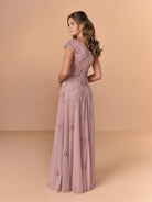 A woman modeling a floor-length dusty pink gown with floral lace embroidery and sheer cap sleeves, shown from the back against a warm beige studio backdrop. / All over beaded gown with sheer cap sleeve, v neckline and full, A-line skirt.