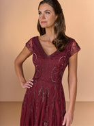 A woman modeling a burgundy lace and mesh A-line dress with intricate sequin embroidery, set against a solid beige studio background. / All over beaded gown with sheer cap sleeve, v neckline and full, A-line skirt.