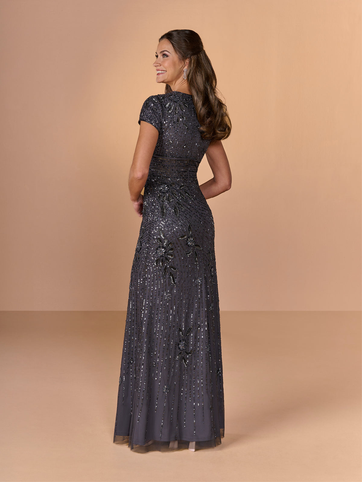 A woman modeling a floor-length evening gown from the back, wearing a dark gray sequined dress with cap sleeves and floral applique, set against a solid beige studio backdrop. / All over beaded gown with v neckline and cap sleeve.