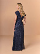 A woman modeling a floor-length navy blue evening gown with sequins and lace, shown from the back against a solid beige studio backdrop. / All over crunchy bead gown with flutter sleeve and cowl back.