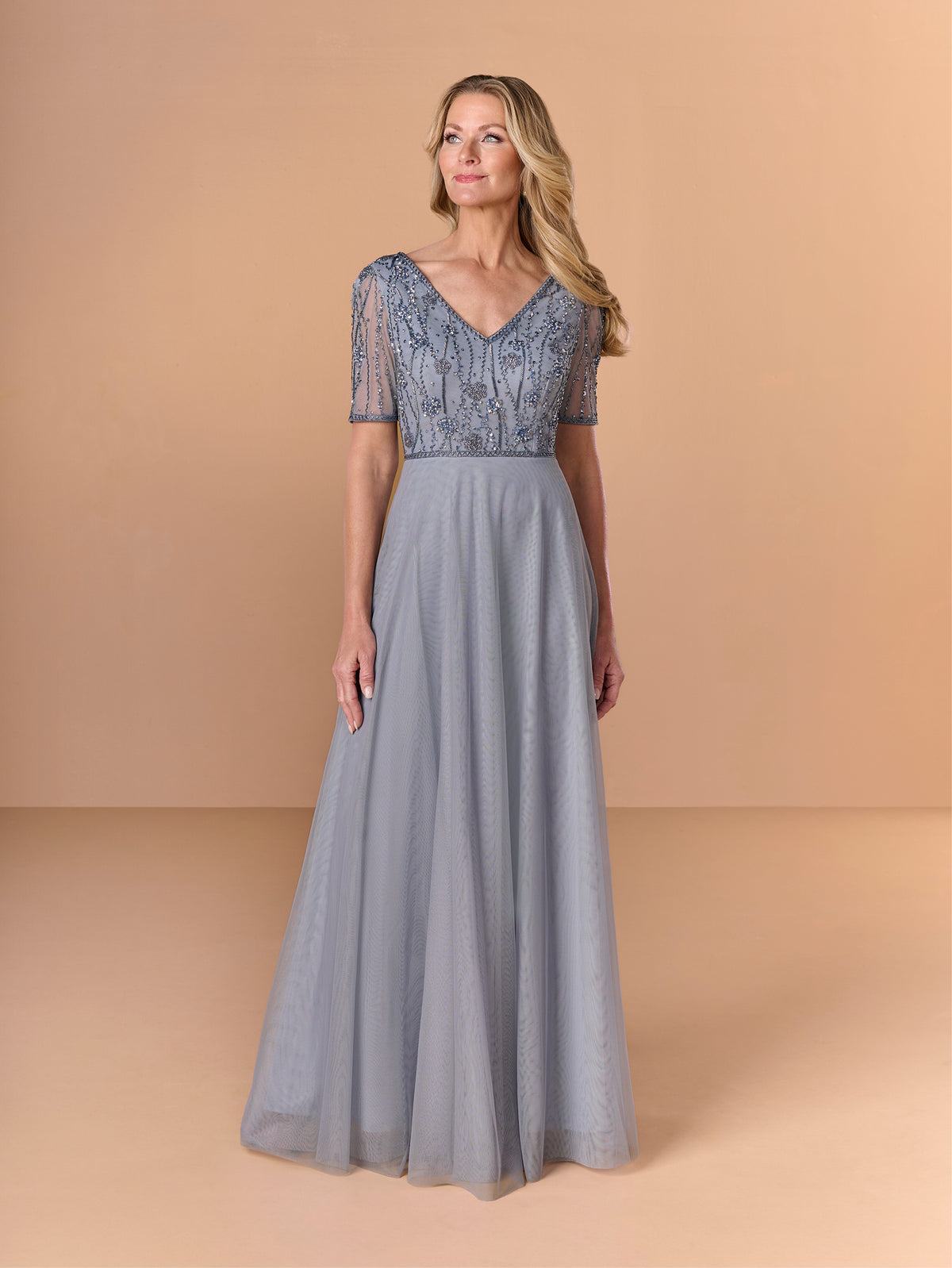 Woman wearing a long, light blue dress with lace top on a beige background / Beaded bodice with v neckline, short sleeve and solid A-line skirt.