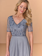 A woman wearing a light blue-gray evening gown with a heavily beaded, sequined V-neck bodice and sheer short sleeves, set against a solid beige studio background. / Beaded bodice with v neckline, short sleeve and solid A-line skirt.