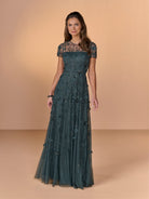 A woman modeling a floor-length teal evening gown with sheer short sleeves, a fitted bodice, and floral applique on a warm beige studio background. / All over beaded gown with illusion neckline and 3D flower detail.