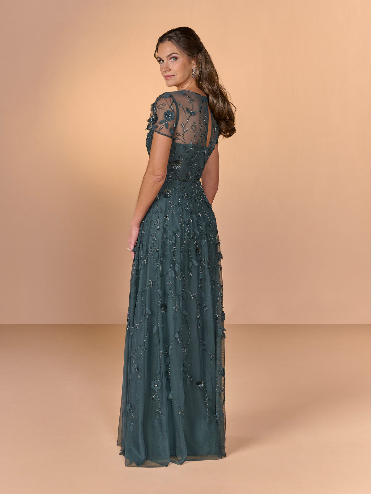 A woman modeling a floor-length teal green evening gown with sheer lace short sleeves and floral applique, shown from the back against a warm beige studio backdrop. / All over beaded gown with illusion neckline and 3D flower detail.
