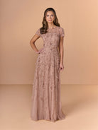 A woman modeling a floor-length dusty rose evening gown with short sleeves, a fitted waist, and floral lace applique, set against a solid beige studio backdrop. / All over beaded gown with illusion neckline and 3D flower detail.
