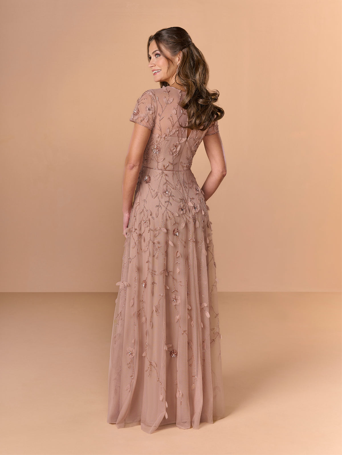 Woman wearing a long, elegant dress with floral embellishments against a beige background / All over beaded gown with illusion neckline and 3D flower detail.