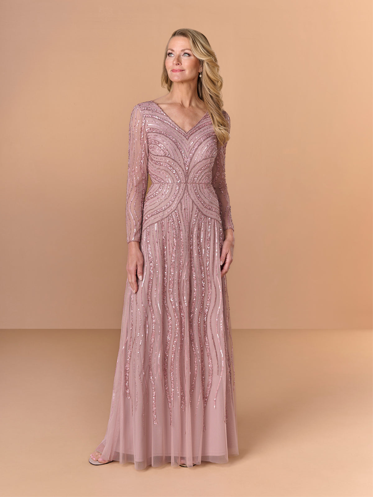 A woman modeling a floor-length, long-sleeve evening gown in a dusty pink color with intricate sequined patterns, set against a solid beige studio backdrop. / All over beaded gown with v neckline, long sleeves and A-line skirt.