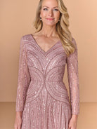 A woman wearing a long-sleeve, V-neck evening gown covered in shimmering rose gold sequins, set against a solid beige studio background. / All over beaded gown with v neckline, long sleeves and A-line skirt.