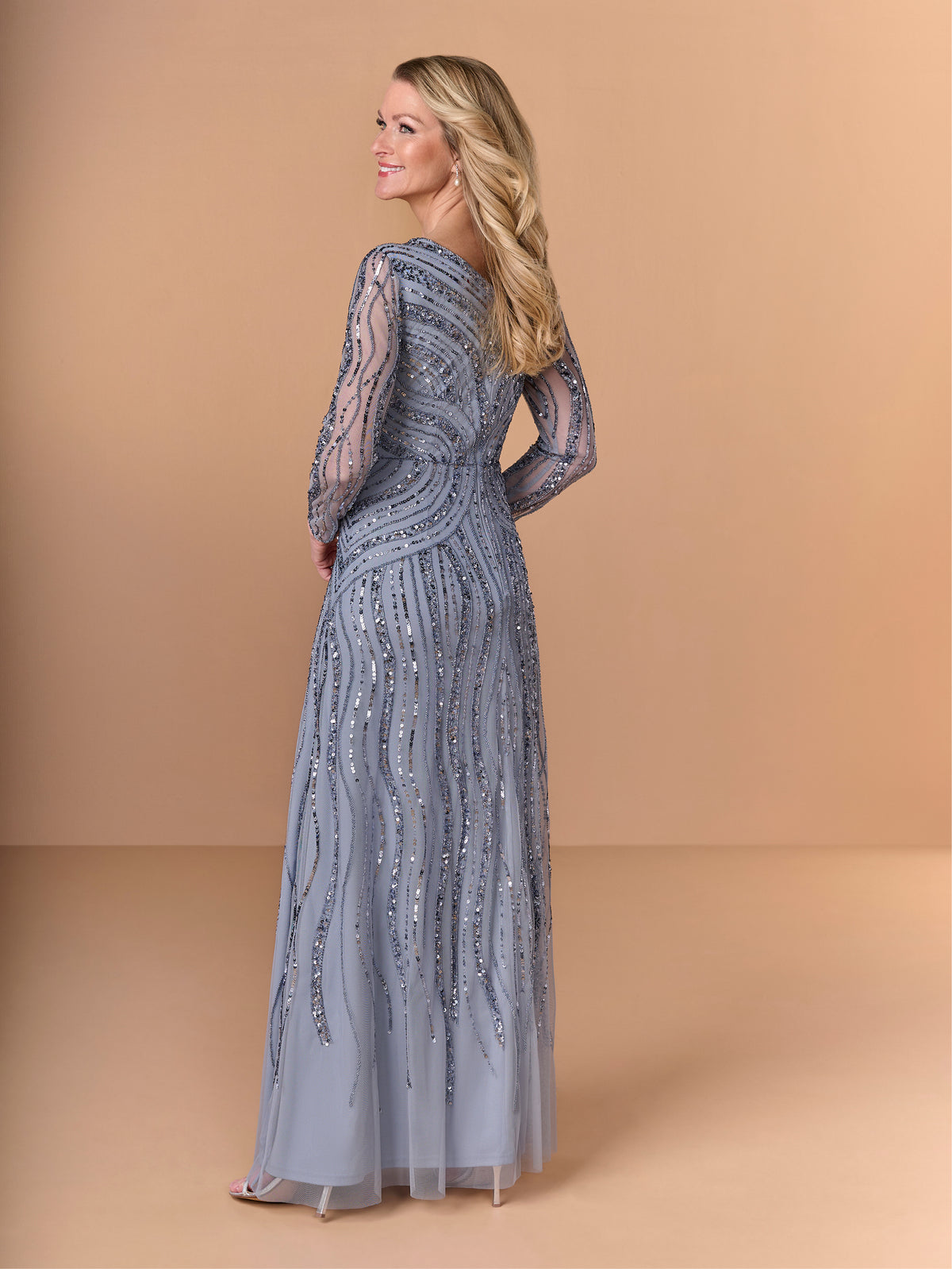 Woman wearing a long, elegant silver dress with lace detailing on a beige background / All over beaded gown with v neckline, long sleeves and A-line skirt.