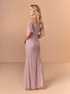 A woman modeling an off-the-shoulder floor-length gown in a dusty rose pink color, featuring intricate beadwork and tulle detailing, set against a solid beige studio background. / All over beaded gown with chic off the shoulder neckline and soft godet skirt.