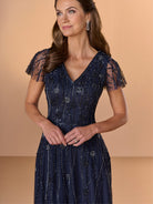 A woman wearing a navy blue evening gown with sheer flutter sleeves, beaded embellishments, and a V-neckline, posed against a solid beige background. / All over beaded gown with v neckline and soft A-line skirt.