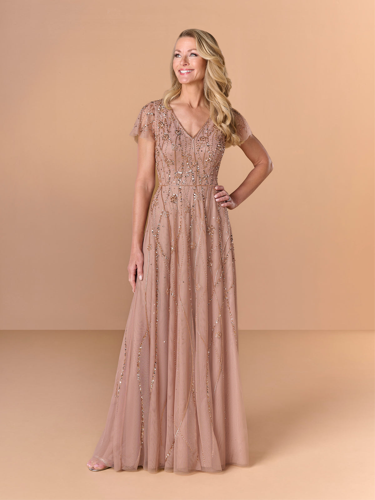 A woman modeling a floor-length, champagne beige evening gown with intricate beadwork and sequin embellishments, set against a solid warm beige studio background. / All over beaded gown with v neckline and soft A-line skirt.