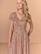 A woman modeling a champagne beige tulle evening gown with intricate gold sequin beadwork and flutter sleeves, set against a solid beige studio background. / All over beaded gown with v neckline and soft A-line skirt.