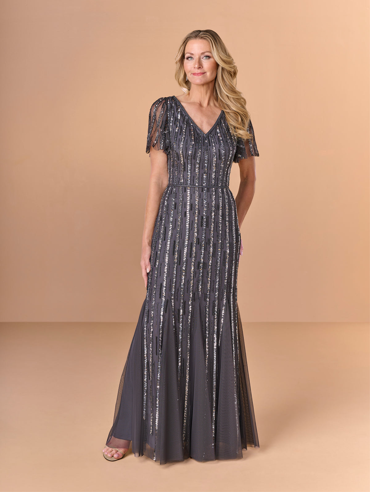 Woman wearing a long, dark gray dress with a subtle pattern on a beige background / All over beaded gown with v neckline, long flutter sleeve and slim, soft godet skirt.