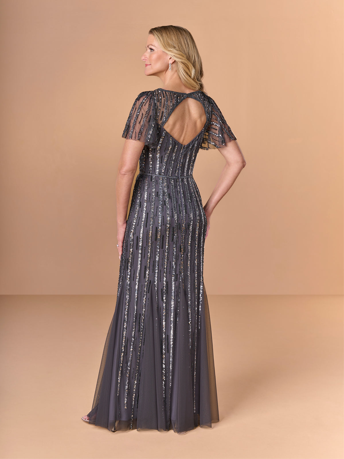 Woman wearing a long, dark blue evening gown with lace details on a beige background / All over beaded gown with v neckline, long flutter sleeve and slim, soft godet skirt.