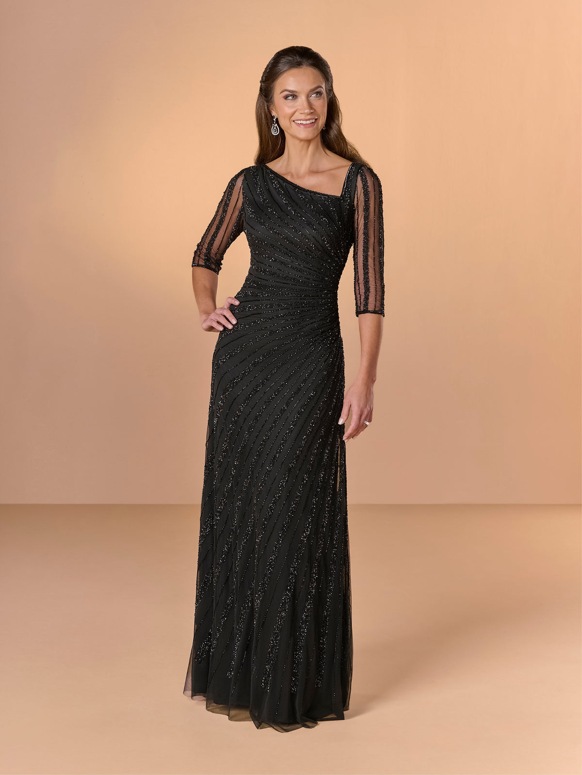 A woman modeling a floor-length black evening gown with sheer three-quarter sleeves and a draped neckline, standing against a warm beige studio backdrop. / All over beaded gown with asymmetrical neckline and body shirring.