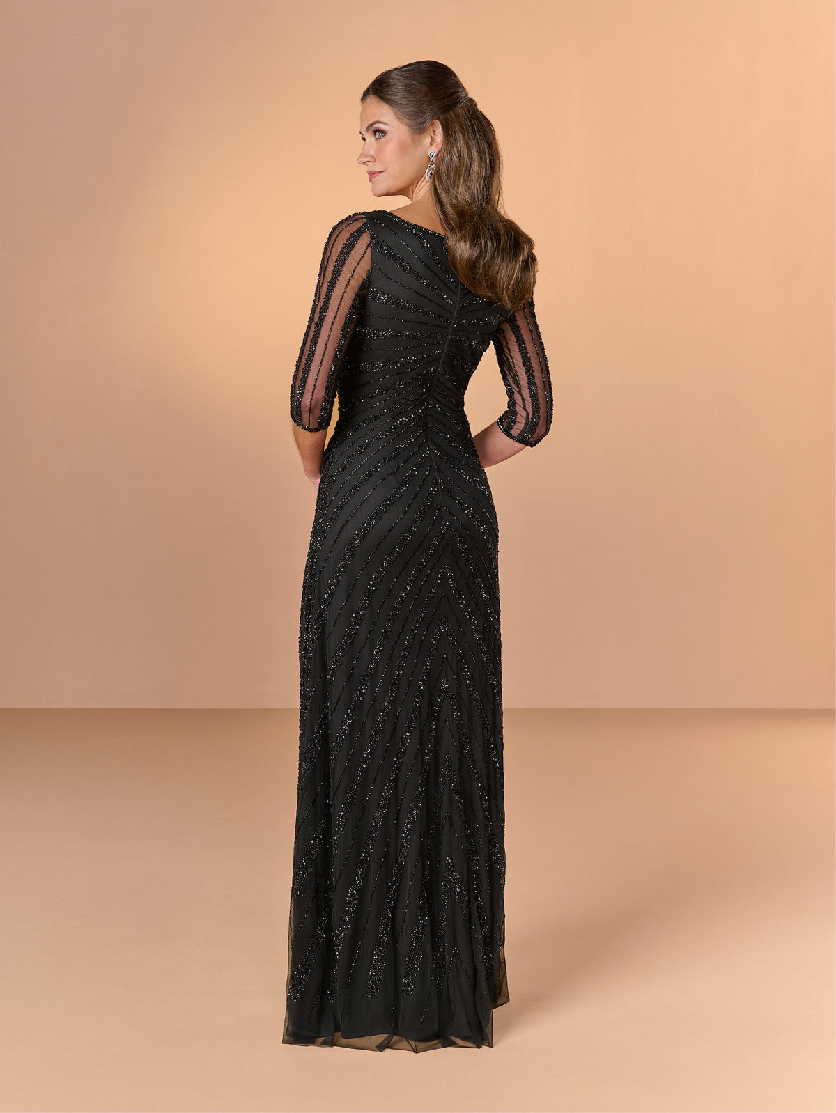 A woman modeling a floor-length black evening gown from the back, featuring sheer three-quarter sleeves and intricate beaded detailing, set against a solid beige studio backdrop. / All over beaded gown with asymmetrical neckline and body shirring.