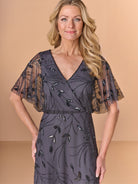 A woman modeling a dark gray wrap-style dress with sheer flutter sleeves and embroidered leaf patterns, posed against a solid beige studio background. / All over beaded gown with surplice, blouson bodice and soft A-line skirt.