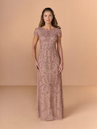 A woman modeling a floor-length, short-sleeve dusty rose gown with intricate floral lace and bead embroidery, set against a solid beige studio backdrop. / All over beaded gown with scoop neck and short sleeve.