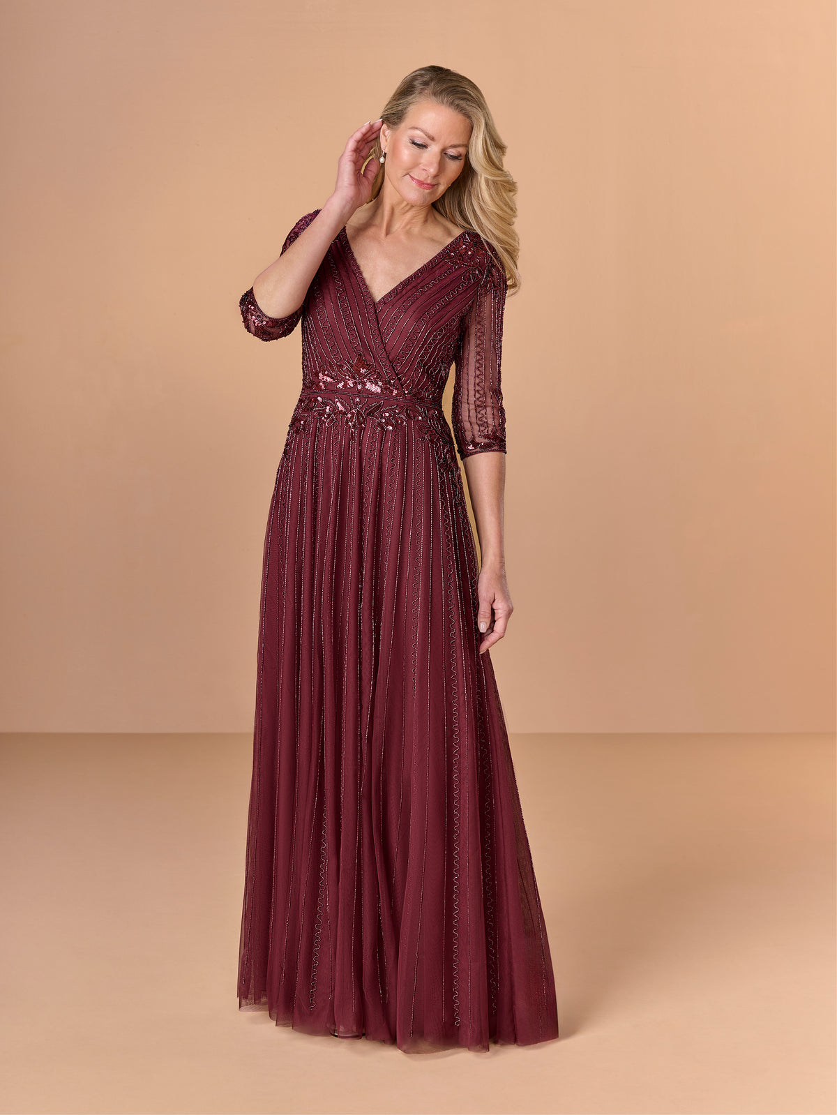 Woman wearing a burgundy evening gown with a beige background / All over beaded gown with surplice bodice, elbow length sleeve and full A-line skirt.