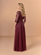 A woman modeling a floor-length burgundy evening gown with three-quarter sleeves, lace detailing, and a pleated tulle skirt, shown from a side-back angle against a solid beige studio backdrop. / All over beaded gown with surplice bodice, elbow length sleeve and full A-line skirt.