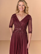 A smiling blonde woman wearing a floor-length burgundy evening gown with sheer three-quarter sleeves, a V-neckline, and intricate beaded vertical detailing, set against a solid beige studio background. / All over beaded gown with surplice bodice, elbow length sleeve and full A-line skirt.