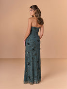 A woman modeling a strapless floor-length gown from the back, shown against a warm beige studio backdrop. The dress is a muted teal-green with black floral embroidery and sequined detailing. / All over beaded gown with high straight neckline, 3D flowers and matching separate shawl.