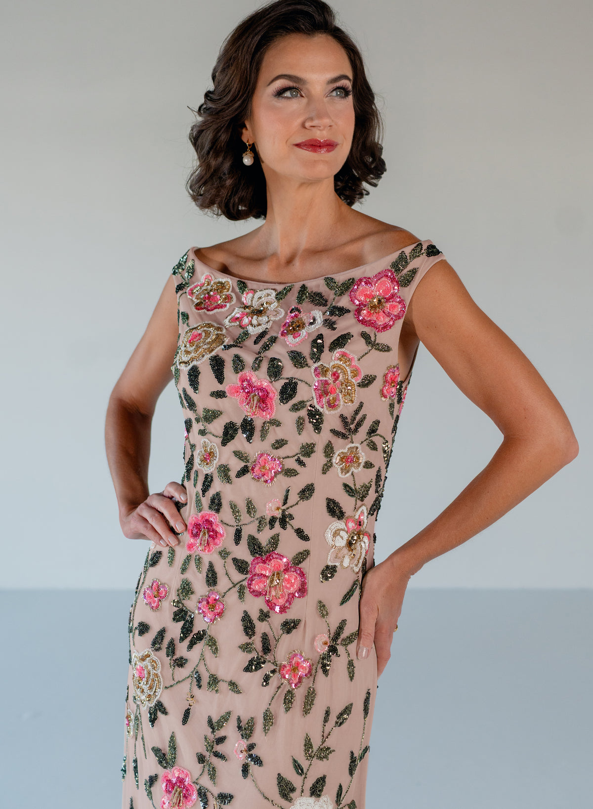 A woman modeling a sleeveless beige dress with pink floral embroidery and green leaf patterns, posed against a neutral light gray background. / Off the shoulder, slim gown with multi color floral beading.