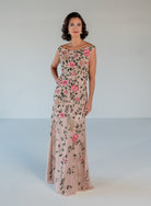 A woman modeling a floor-length off-the-shoulder gown in blush pink with an all-over floral and vine embroidery pattern in pink, green, and gold, set against a plain light gray studio background. / Off the shoulder, slim gown with multi color floral beading.