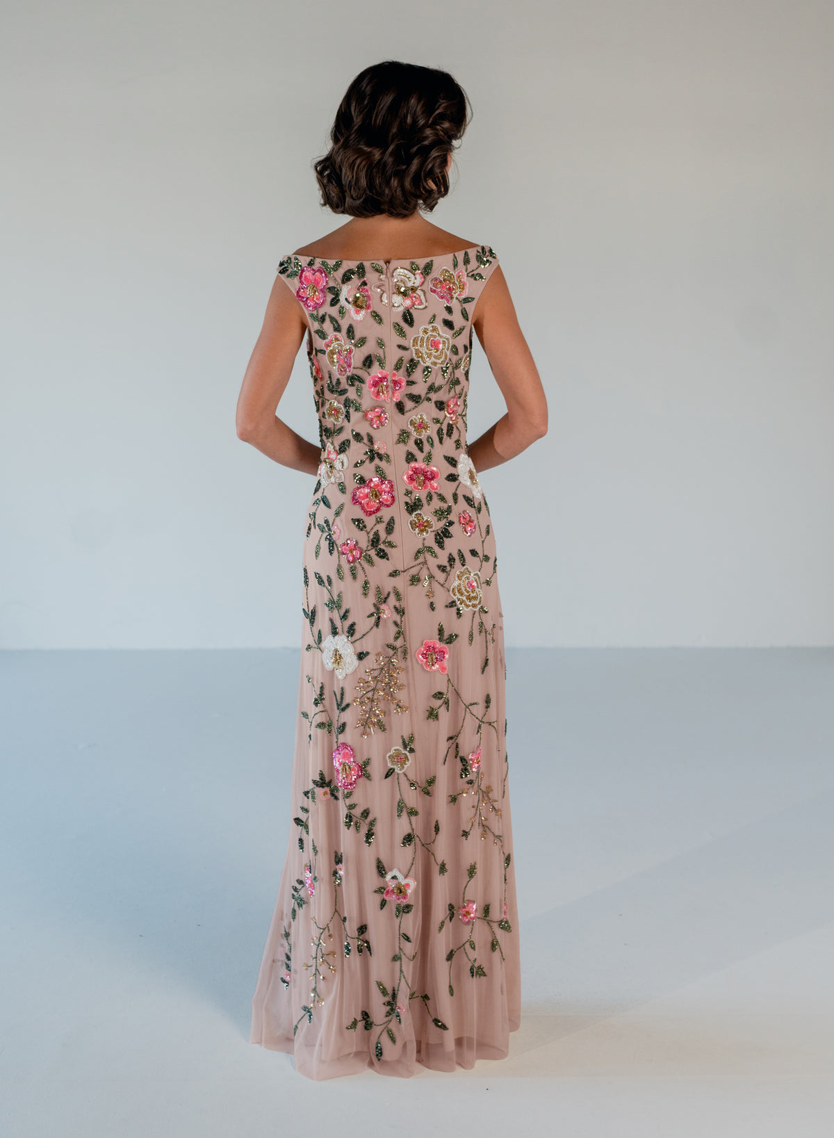 A woman viewed from behind wearing a floor-length blush pink gown with floral embroidery and tulle detailing, standing against a plain light gray studio background. / Off the shoulder, slim gown with multi color floral beading.