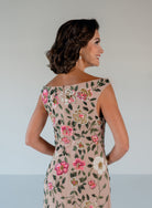 A woman shown from the back wearing a fitted off-the-shoulder dress in blush beige with colorful floral embroidery in pink, green, and gold, against a light gray studio background. / Off the shoulder, slim gown with multi color floral beading.
