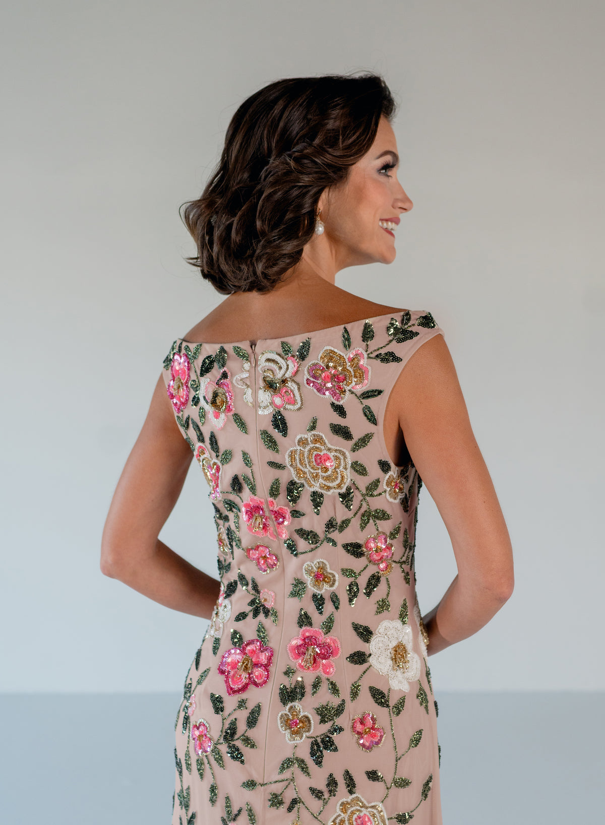 A woman shown from the back wearing a fitted off-the-shoulder dress in blush beige with colorful floral embroidery in pink, green, and gold, against a light gray studio background. / Off the shoulder, slim gown with multi color floral beading.