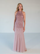 A woman modeling a sleeveless, high-neck, floor-length gown in a dusty pink color with dense beaded embellishments across the bodice and a flowing tulle skirt, set against a plain light gray studio background. / Halter neckline slim gown with soft godets and all over ombre beading.