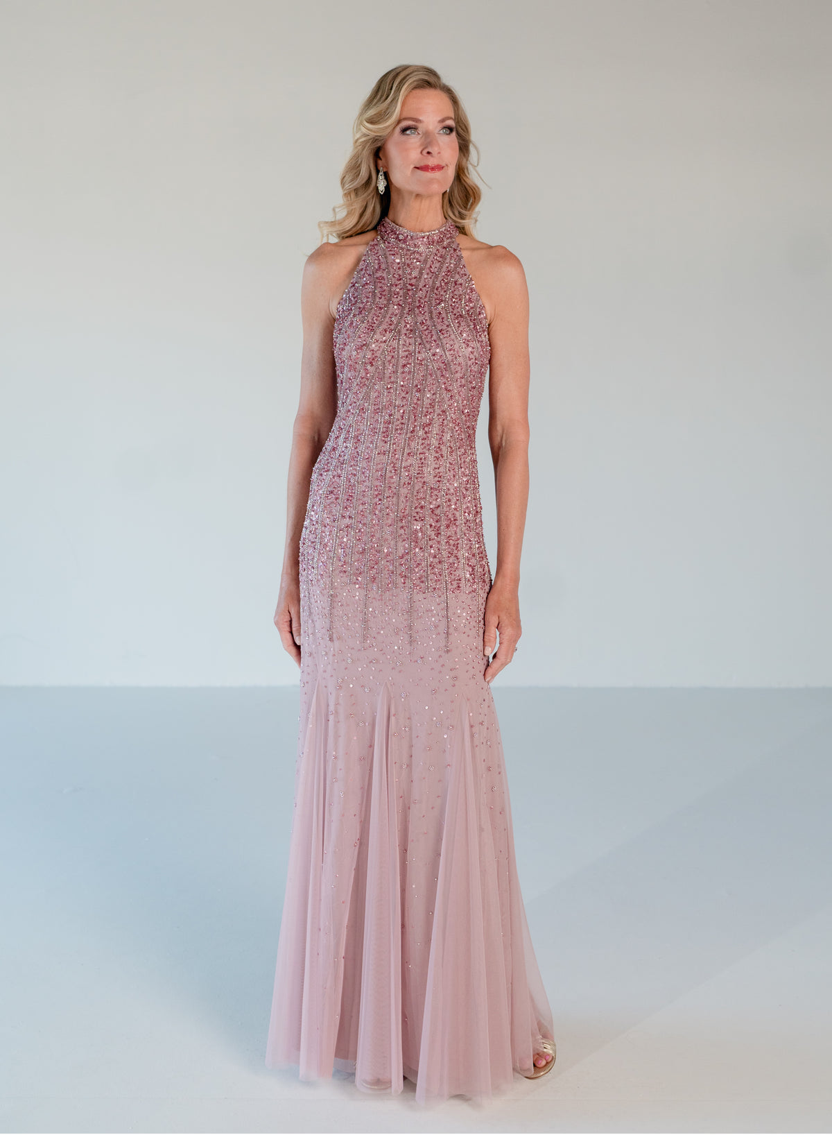 A woman modeling a sleeveless, high-neck, floor-length gown in a dusty pink color with dense beaded embellishments across the bodice and a flowing tulle skirt, set against a plain light gray studio background. / Halter neckline slim gown with soft godets and all over ombre beading.
