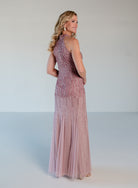 A woman modeling a sleeveless, high-neck, floor-length gown with dense beaded embellishment and a flowing tulle skirt, photographed against a plain light gray studio background. / Halter neckline slim gown with soft godets and all over ombre beading.