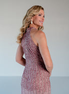A woman with blonde wavy hair is shown from the back wearing a sleeveless, high-neck, floor-length evening gown covered in shimmering pink and silver sequins, set against a plain light gray studio background. / Halter neckline slim gown with soft godets and all over ombre beading.
