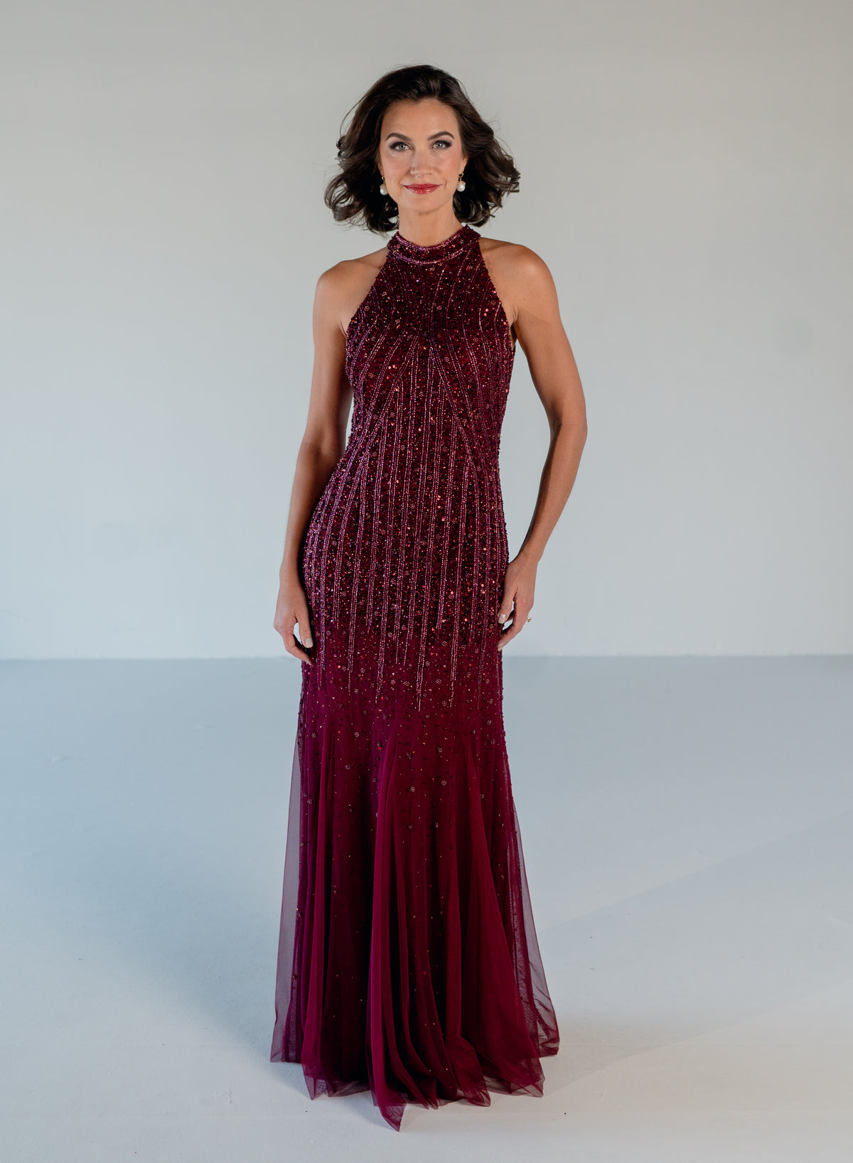 A woman modeling a sleeveless, high-neck floor-length gown in deep burgundy with dense vertical beadwork and sequins, set against a plain light gray studio background. / Halter neckline slim gown with soft godets and all over ombre beading.