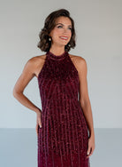 A woman modeling a sleeveless, high-neck halter gown covered in shimmering dark burgundy sequins, set against a plain light gray studio background. / Halter neckline slim gown with soft godets and all over ombre beading.
