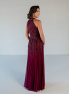 A woman modeling a sleeveless, high-neck floor-length gown with a fitted bodice covered in vertical beaded stripes and a flowing tulle skirt, shown from the back against a plain light gray studio background. / Halter neckline slim gown with soft godets and all over ombre beading.