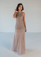 A woman modeling a sleeveless, high-neck floor-length gown covered in shimmering sequins and beadwork, with a flowing tulle skirt, against a plain light gray studio background. / Halter neckline slim gown with soft godets and all over ombre beading.