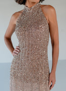 A sleeveless halter-neck evening gown covered in dense vertical beading and sequins, shown on a model against a light gray studio background. / Halter neckline slim gown with soft godets and all over ombre beading.