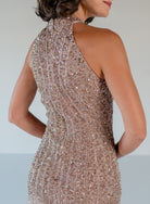 A woman shown from the back wearing a sleeveless, high-neck, bodycon dress covered in dense metallic rose-gold sequins and beadwork against a plain light gray studio background. / Halter neckline slim gown with soft godets and all over ombre beading.