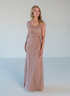 A woman modeling a floor-length, sleeveless evening gown in a dusty rose pink color with shimmering sequin and bead embellishments, set against a plain light gray studio background. / V neckline all over beaded gown with shirring and short sleeves.