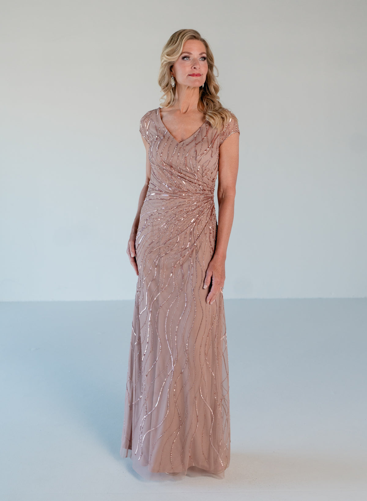 A woman modeling a floor-length, sleeveless evening gown in a dusty rose pink color with shimmering sequin and bead embellishments, set against a plain light gray studio background. / V neckline all over beaded gown with shirring and short sleeves.