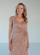 A woman modeling a sleeveless, V-neck evening gown covered in shimmering rose-gold sequins and beadwork, set against a clean light gray studio background. / V neckline all over beaded gown with shirring and short sleeves.