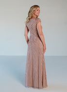 A woman modeling a floor-length evening gown from the back, featuring a dusty rose pink color with intricate bead and sequin embellishments, set against a plain light gray studio background. / V neckline all over beaded gown with shirring and short sleeves.
