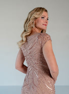 A woman with blonde wavy hair is shown from the back wearing a sleeveless, floor-length evening gown covered in shimmering rose-gold sequins and beadwork against a plain light gray studio background. / V neckline all over beaded gown with shirring and short sleeves.