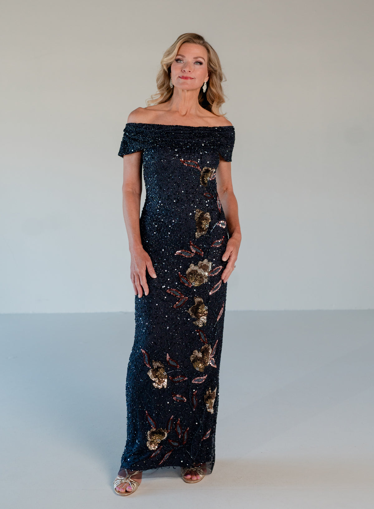 A woman modeling an off-the-shoulder, floor-length navy gown covered in shimmering sequins and decorated with large metallic gold and bronze floral motifs, set against a plain light gray studio background. / Off the shoulder column gown with all over beading and multi color detailing.