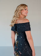 A woman with blonde wavy hair is shown from the back wearing a dark navy off-the-shoulder sequined dress with gold and red floral accents, set against a plain light gray background. / Off the shoulder column gown with all over beading and multi color detailing.