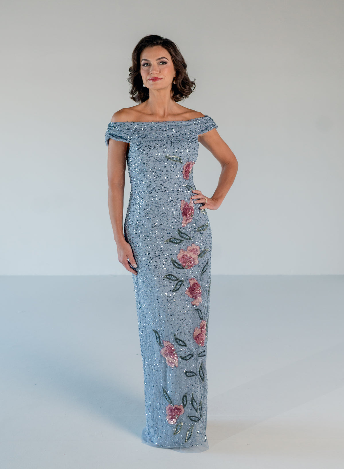 A woman modeling an off-the-shoulder floor-length gown covered in light blue sequins with pink floral and green leaf embroidery, set against a plain light gray studio background. / Off the shoulder column gown with all over beading and multi color detailing.
